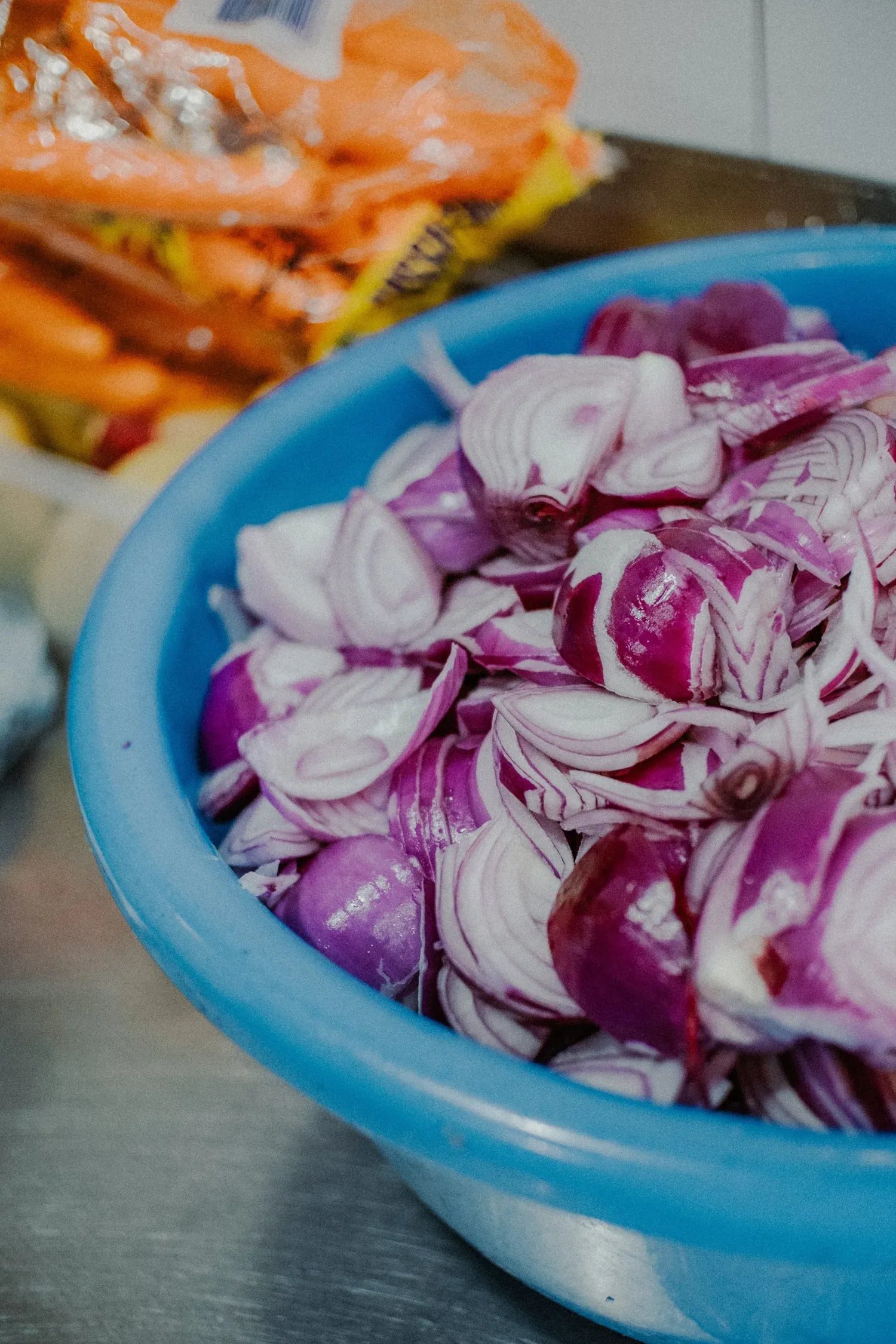 Blue bowl filled with freshly chopped red onions on a metal surface with vegetables in background.