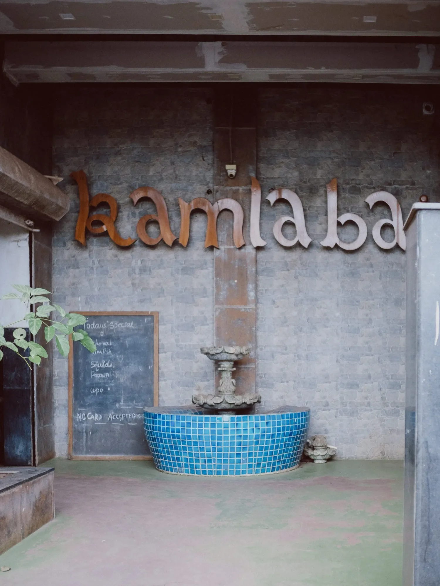 Kamla Bar entrance with rustic metal lettering on concrete wall and blue mosaic tiled fountain.