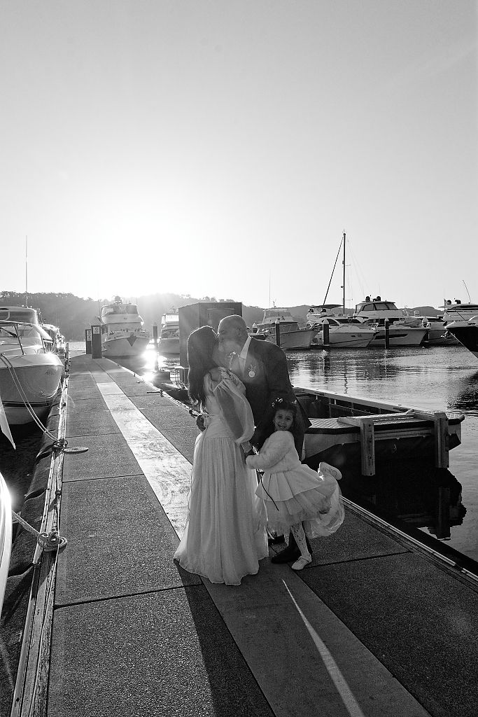 Black and white photo of a couple kissing on a marina dock at sunset while a young girl in a dress stands beside them near docked boats.