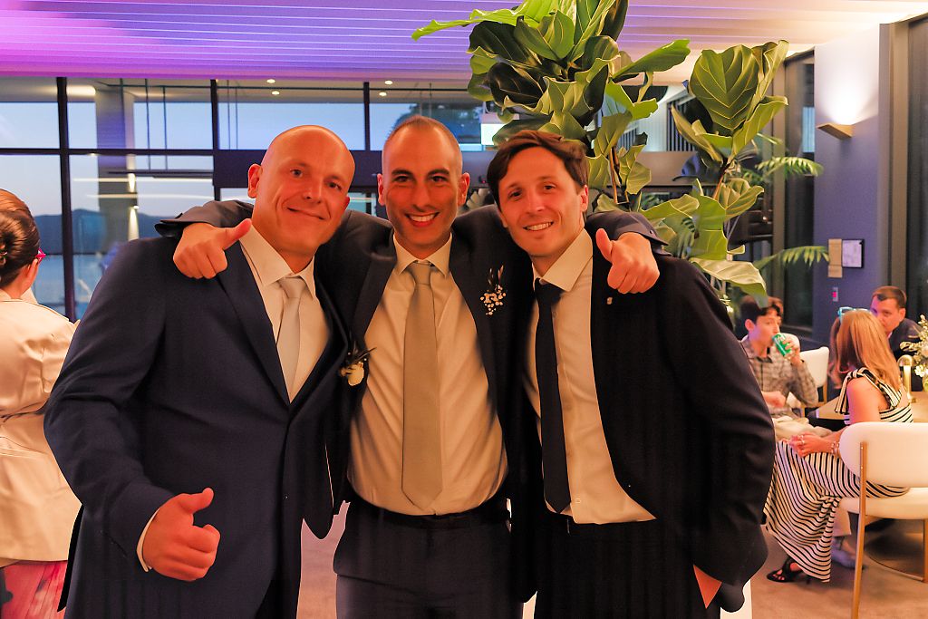 Three smiling men in dark jackets pose with arms around each other at an indoor celebration with large green plants behind them.