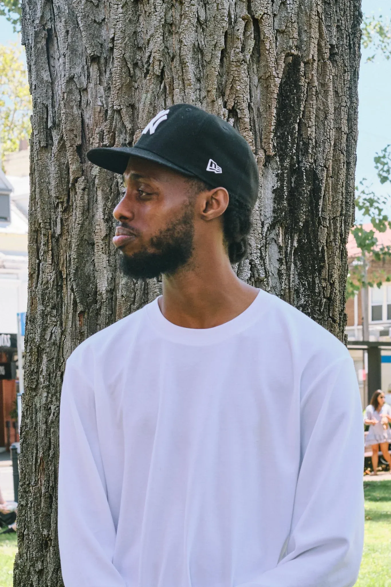 Black man in white long sleeve shirt and NY Yankees cap leaning against tree trunk outdoors.