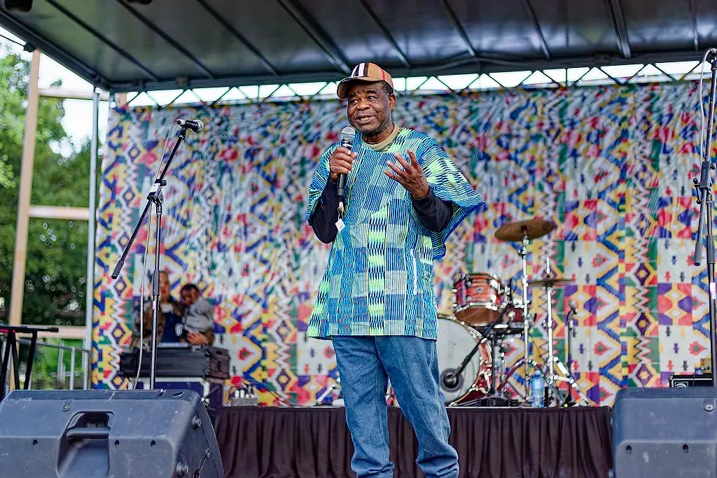 Man in blue African print shirt speaking into microphone on outdoor festival stage.