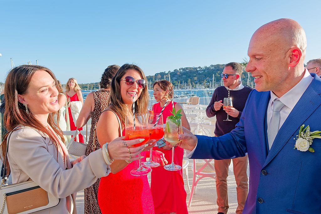 Smiling wedding guests raise colorful cocktails in a toast with the groom on a sunny waterfront terrace.
