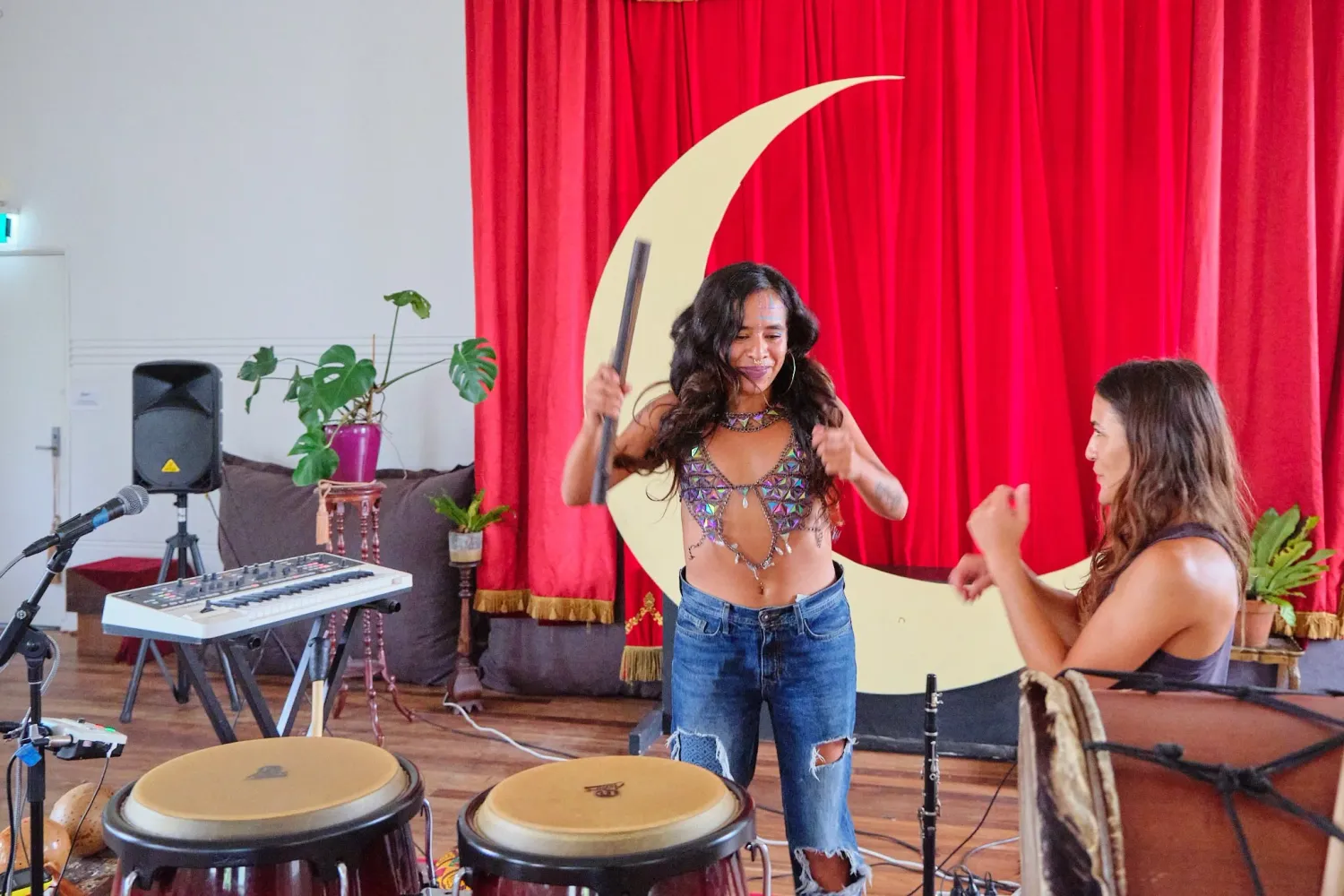 Two women perform together with drums in a bohemian studio with red curtains and crescent moon decor.