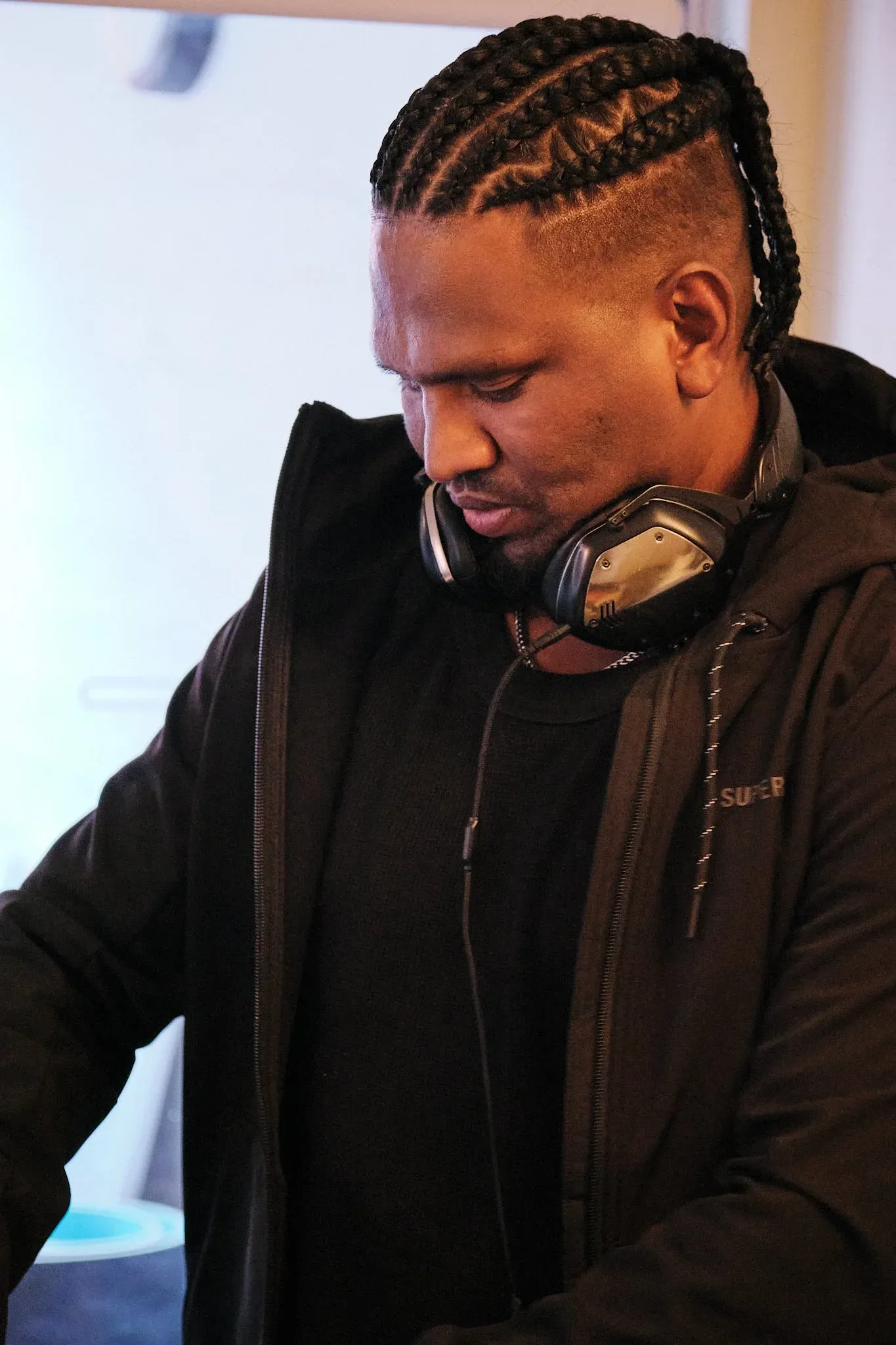 Man with cornrow braids wearing black hoodie and headphones around neck, looking down at DJ equipment.