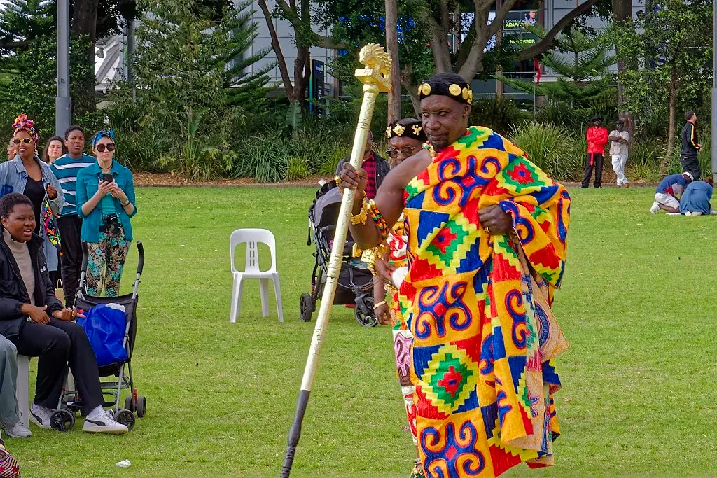 Performer in traditional colorful kente cloth regalia carrying a ceremonial staff at an African festival.