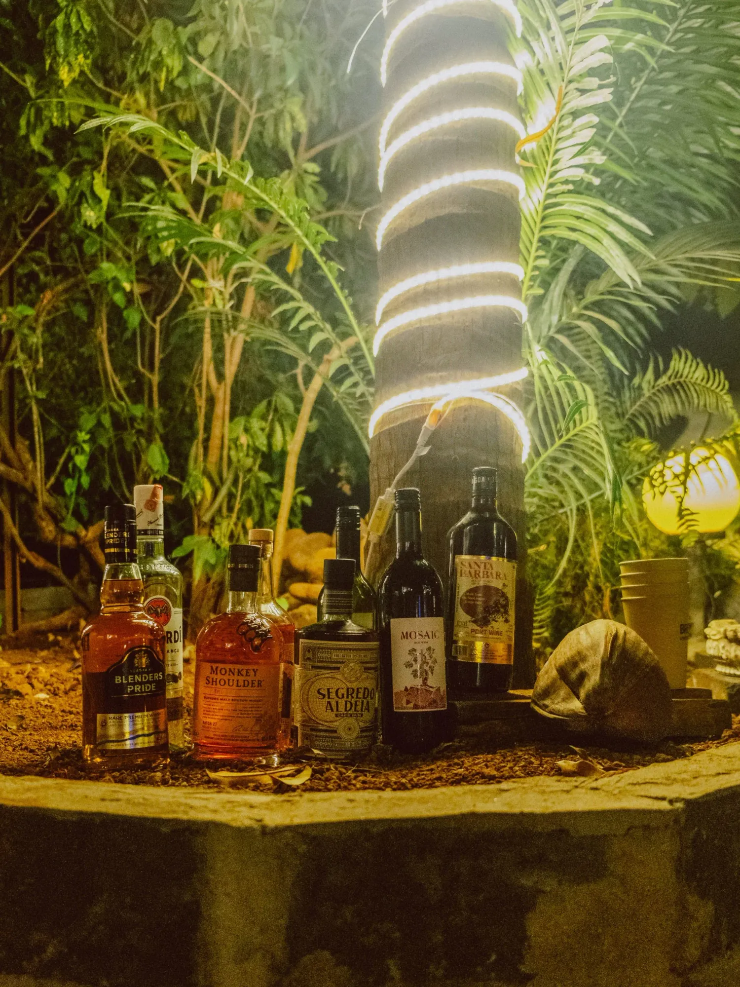 Collection of whiskey and spirits bottles arranged outdoors under spiral neon light amid tropical foliage.