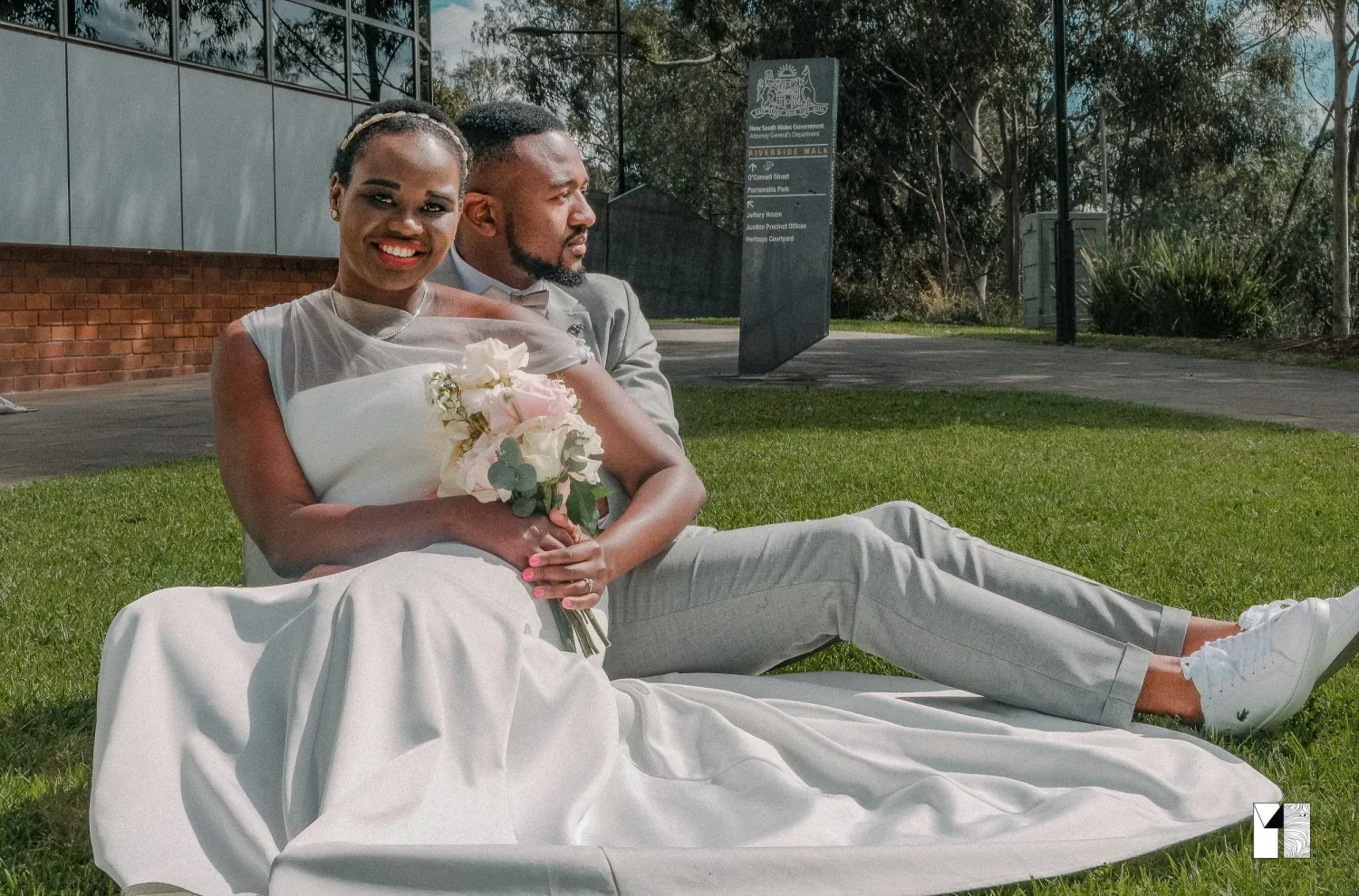 Bride and groom sitting on grass, bride in white gown holding pink bouquet, groom in gray suit.