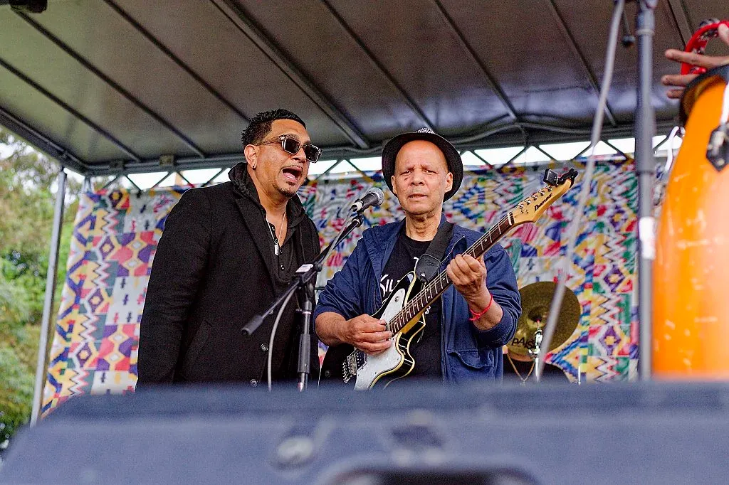Two male musicians perform together on stage, one singing and one playing an electric guitar.
