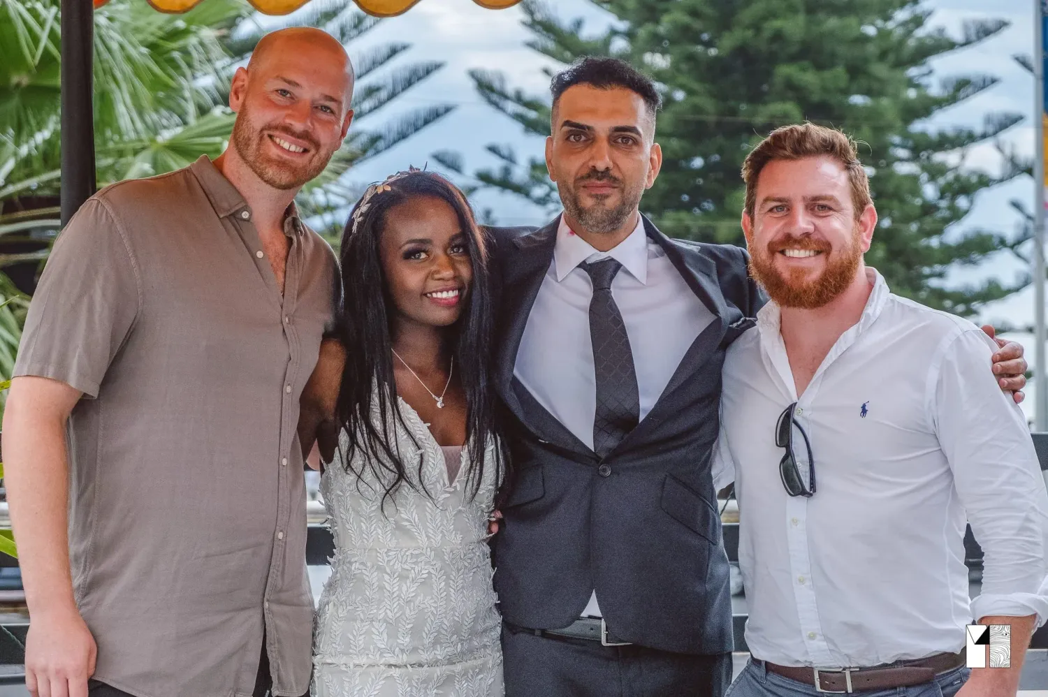 Happy couple celebrates with two male friends at sunny outdoor wedding reception.