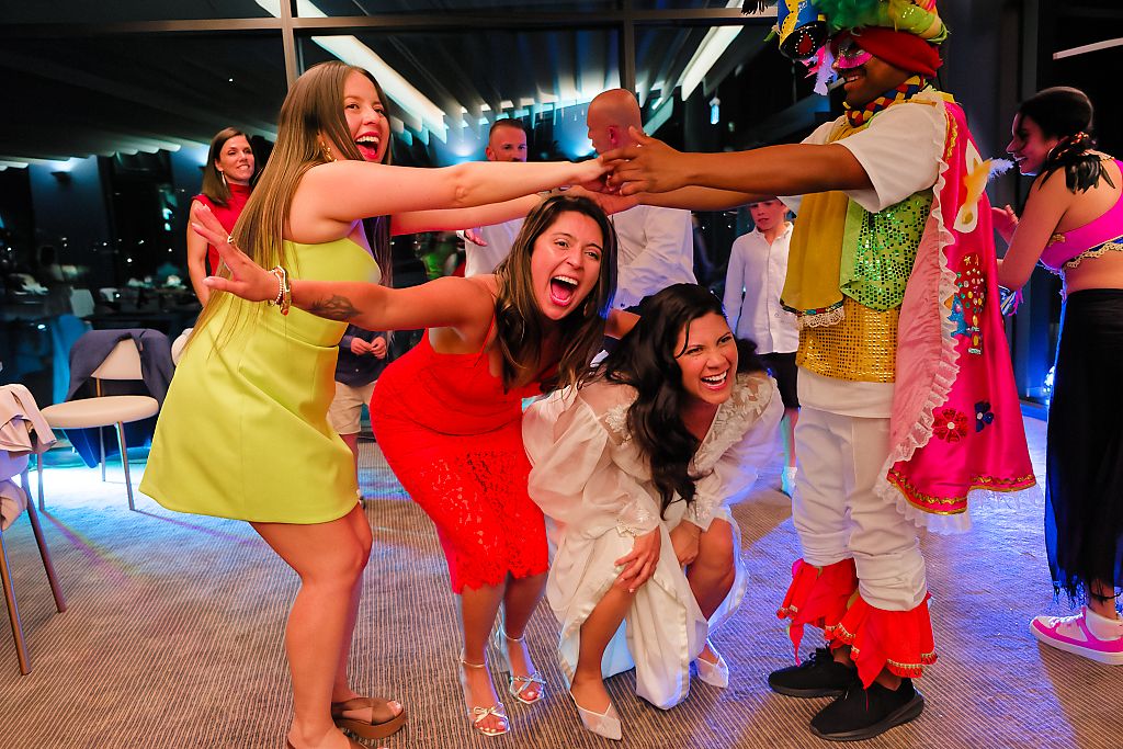 Guests in colorful dresses laugh and dance together with a performer in a vibrant festive costume at a lively evening celebration.