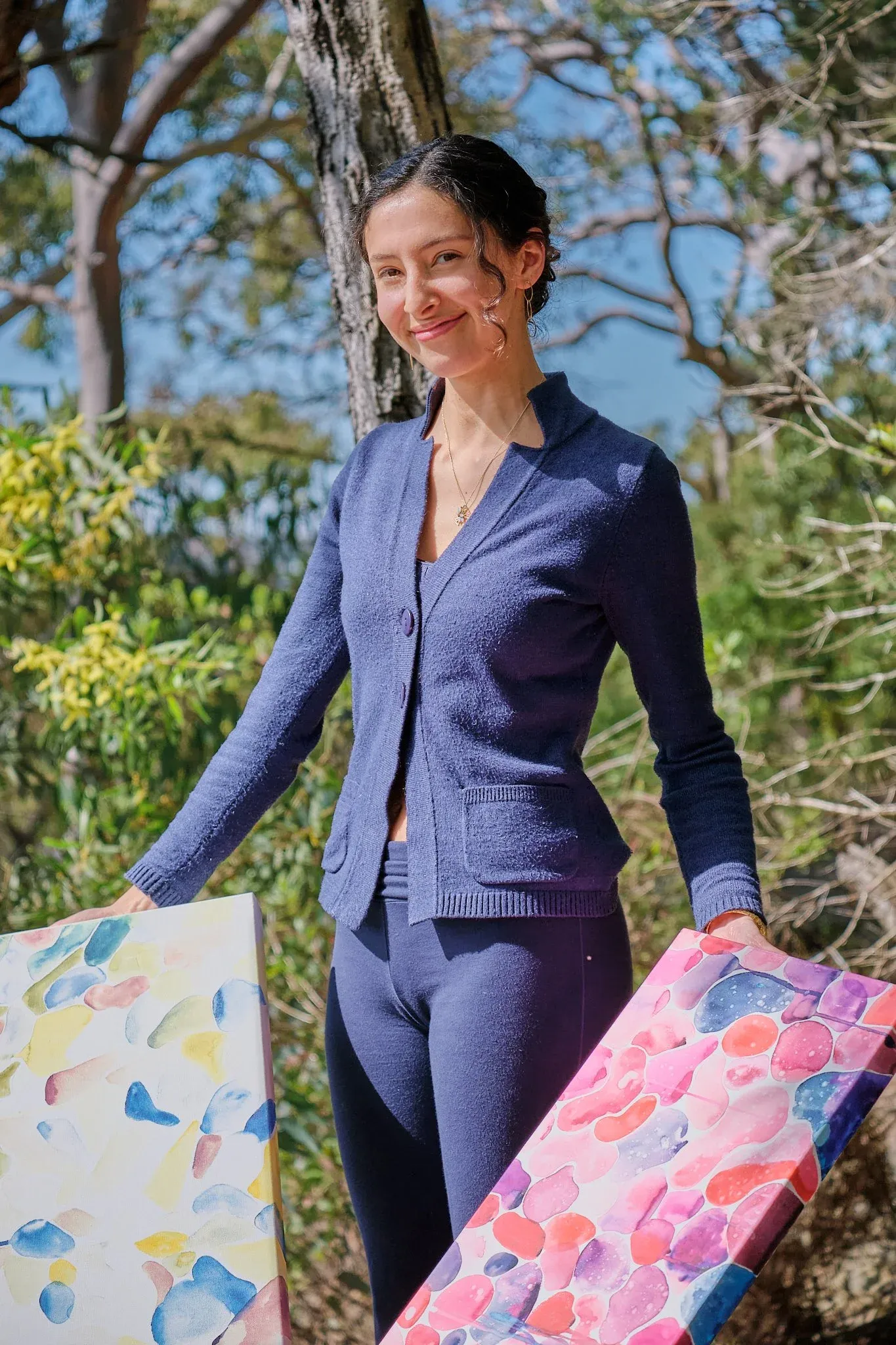 Woman in blue knit suit holding colorful abstract paintings outdoors among trees.