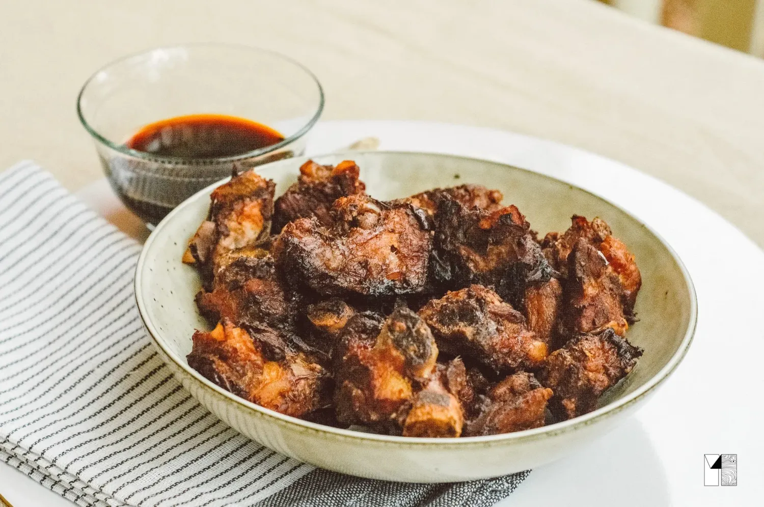 Bowl of crispy fried pork pieces with a side of dark dipping sauce on a striped cloth.