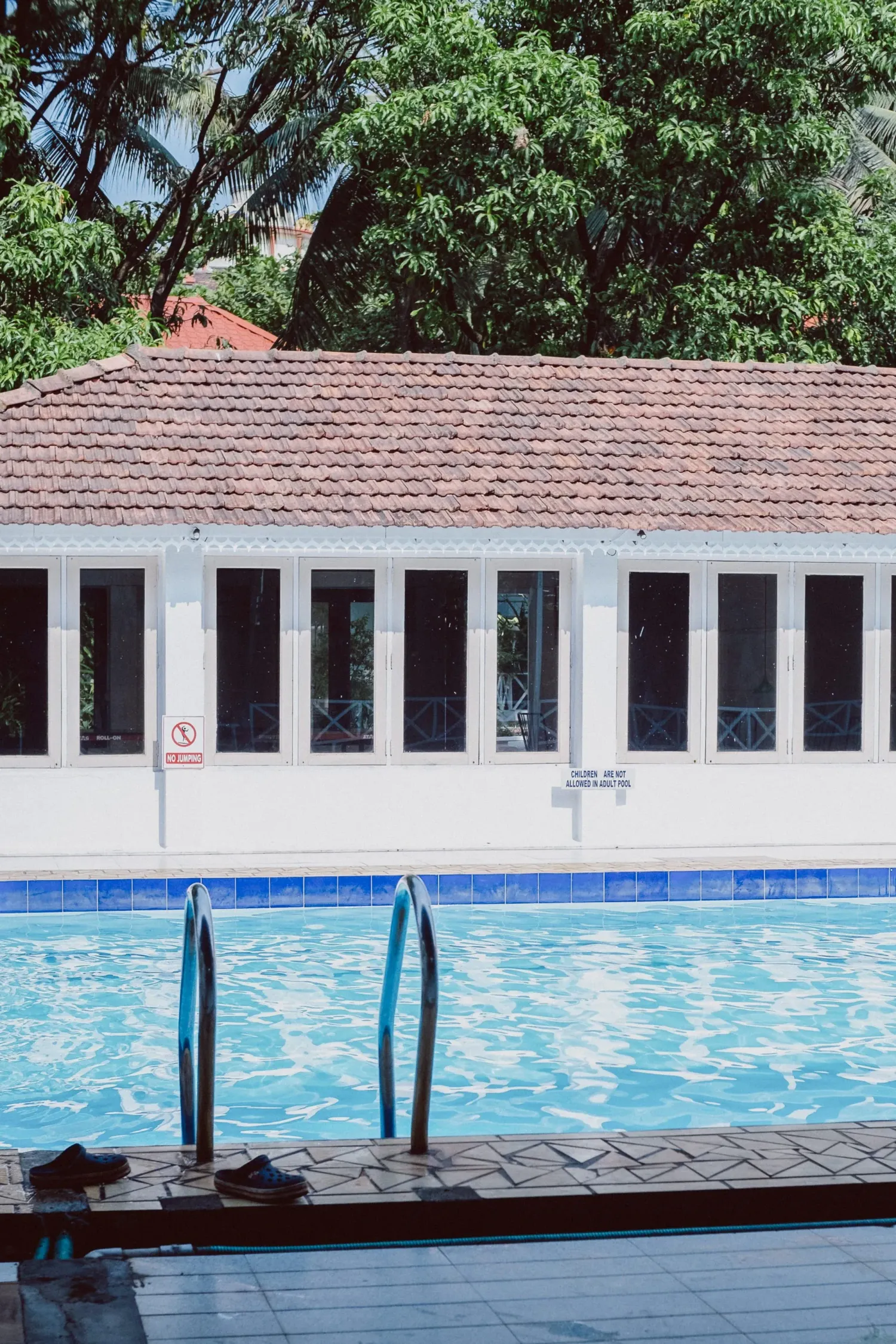 Outdoor swimming pool with metal handrails beside white colonial building surrounded by lush green trees.
