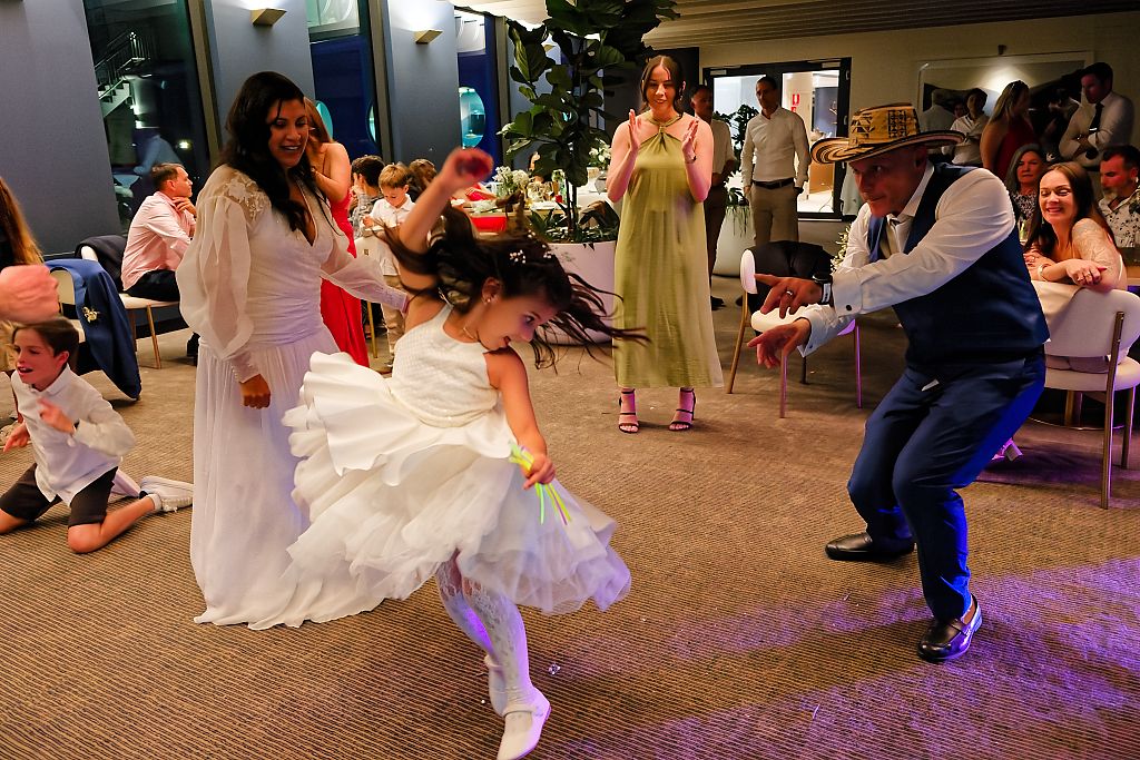Young girl in a white tutu dress twirls on the dance floor as guests, including a man in a hat, dance and watch at a celebration.