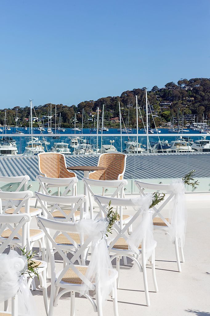 White wedding ceremony chairs with tulle bows and greenery on a sunny waterfront terrace overlooking a marina filled with sailboats.