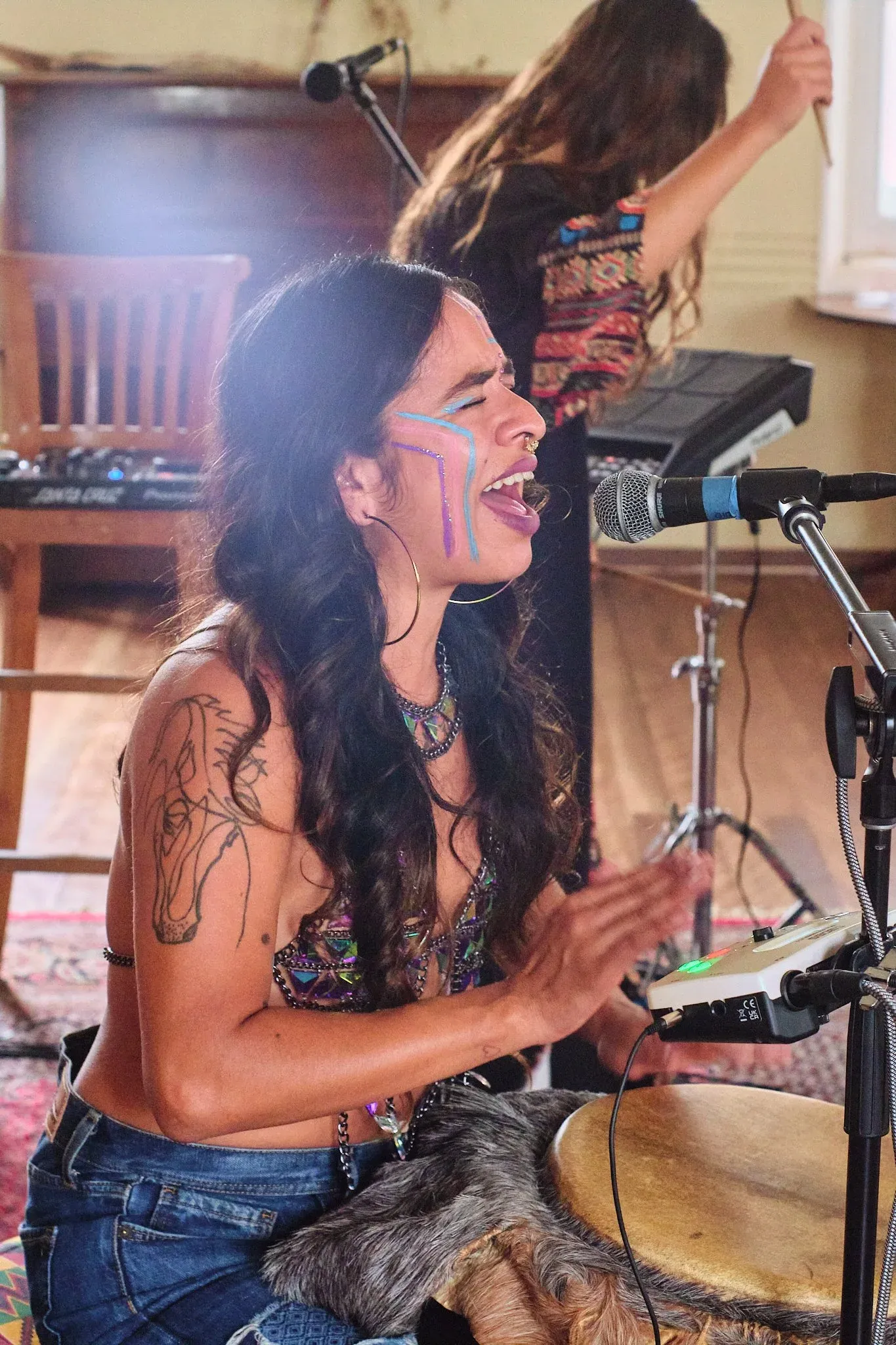 Woman with face paint plays djembe drum and sings into microphone at an indoor music performance.