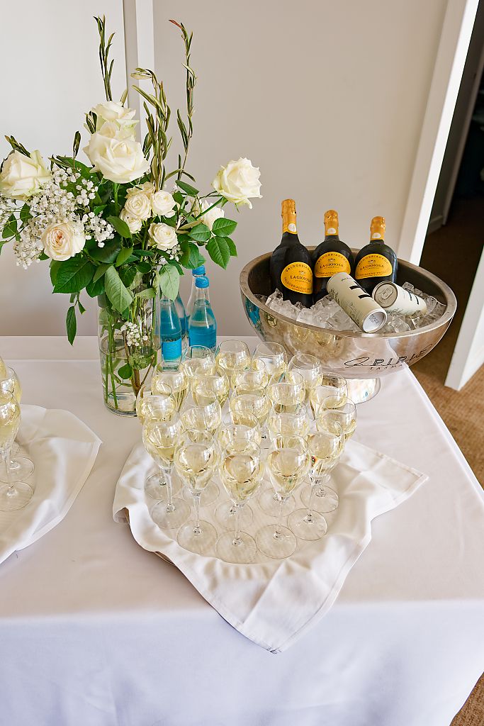 Champagne flutes on a white linen table beside bottles chilling in a silver ice bucket and a vase of white roses.