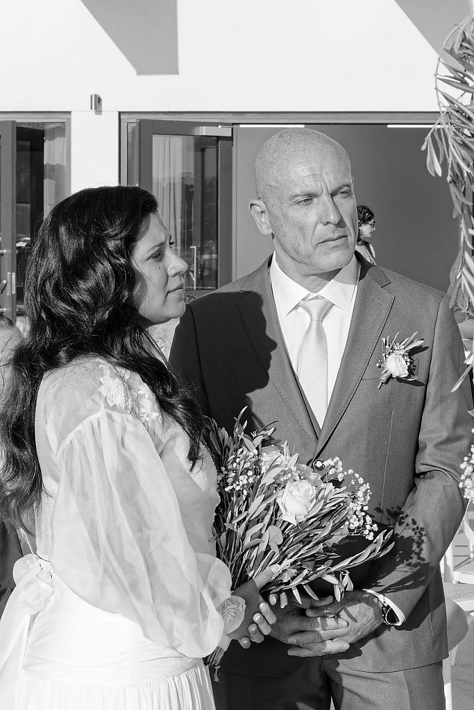 Black and white photo of a bride in a lace dress holding a bouquet beside a bald groom in a suit with a boutonniere at a wedding ceremony.