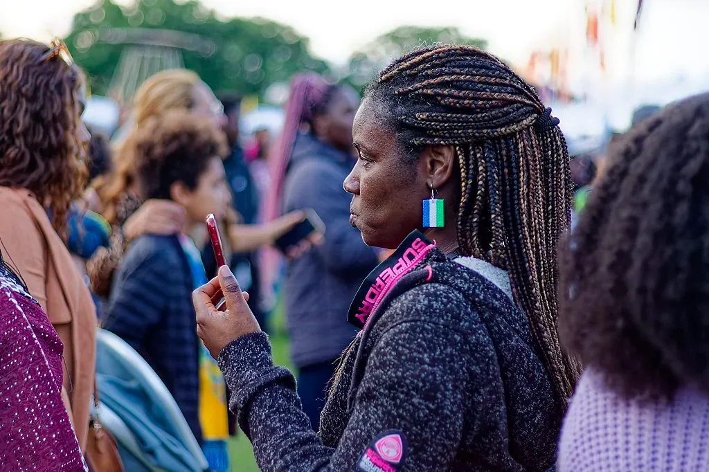 Woman with long braids and colorful earrings using smartphone at crowded outdoor cultural festival.