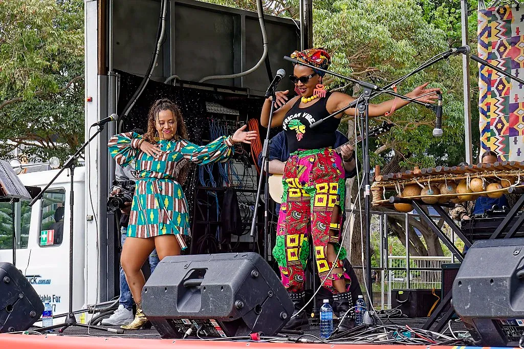 Two women performing on outdoor stage wearing vibrant African print clothing with microphones.