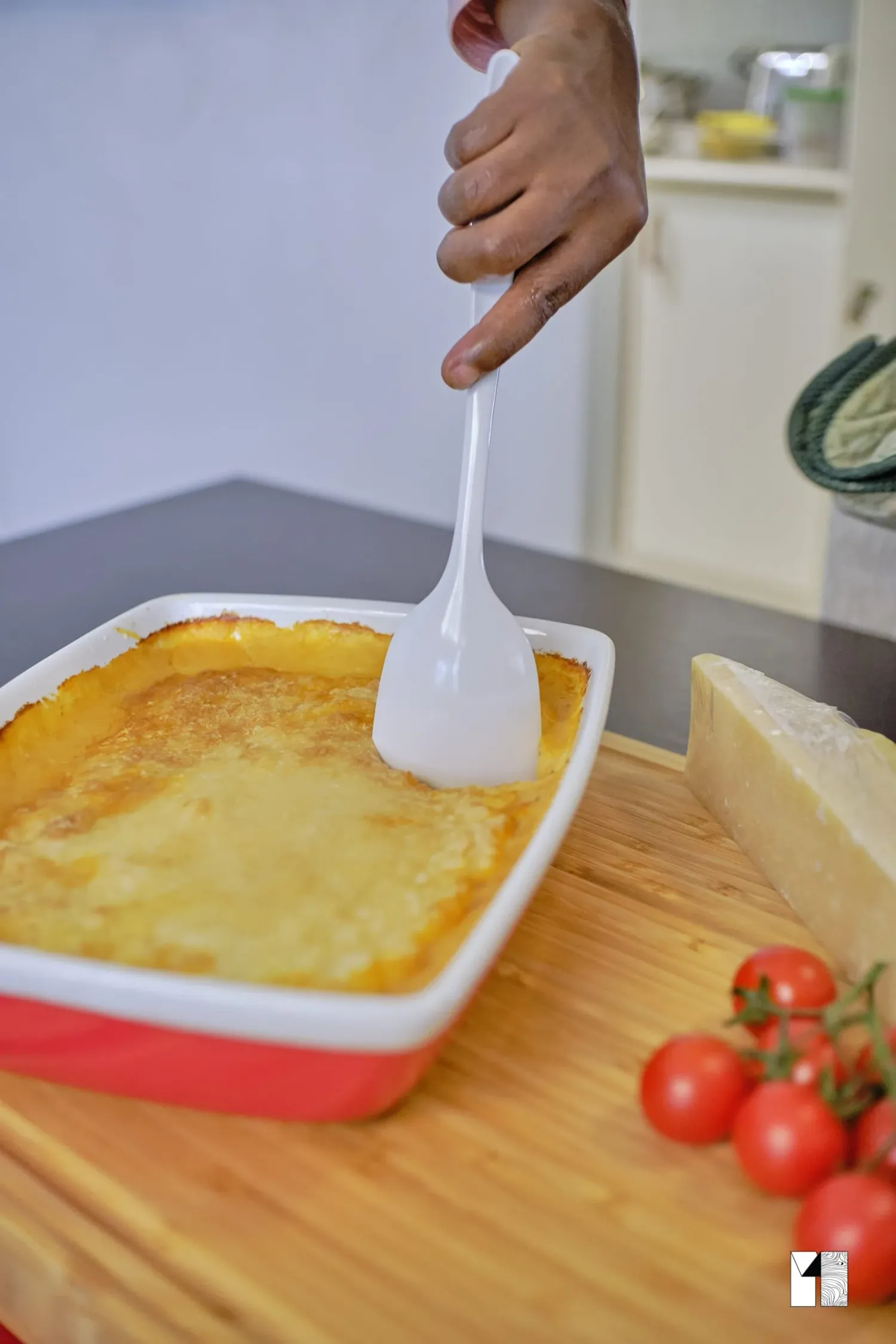 Hand serving cheesy baked lasagna from a red and white dish on a wooden board with tomatoes.