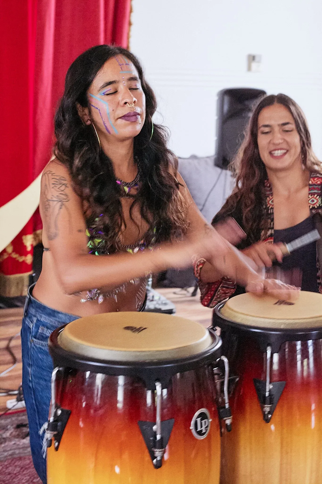 Face-painted woman energetically playing LP conga drums with another female musician in background.