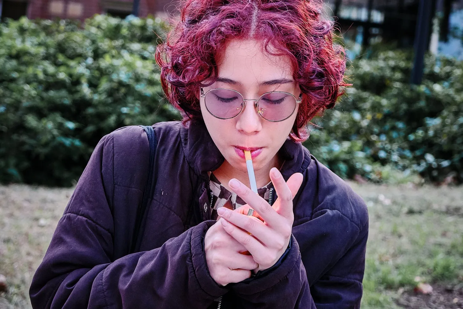 Young woman with red curly hair lighting a cigarette outdoors in a park.