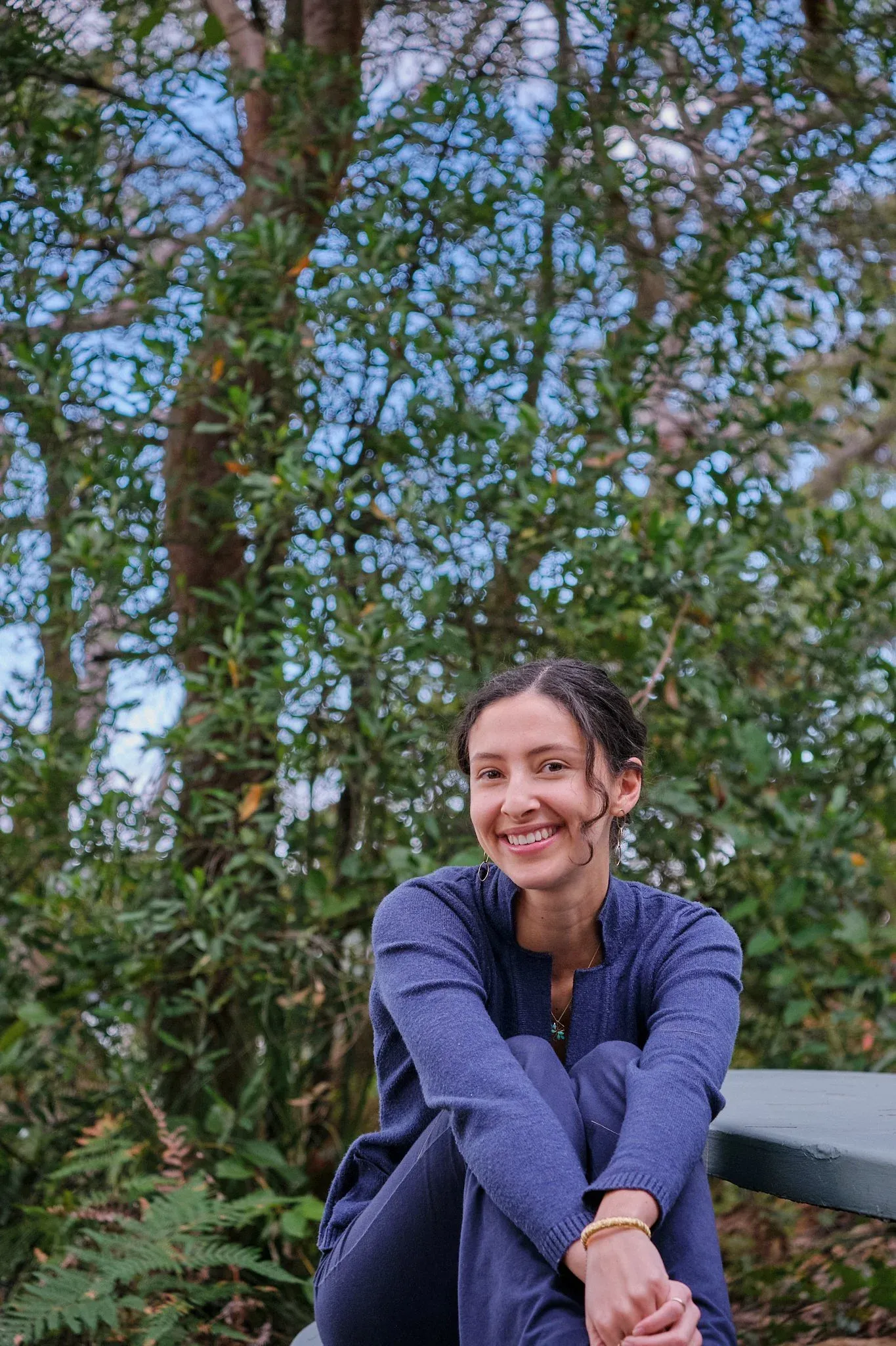 Smiling woman in blue outfit sitting on a bench surrounded by lush green Australian bushland.