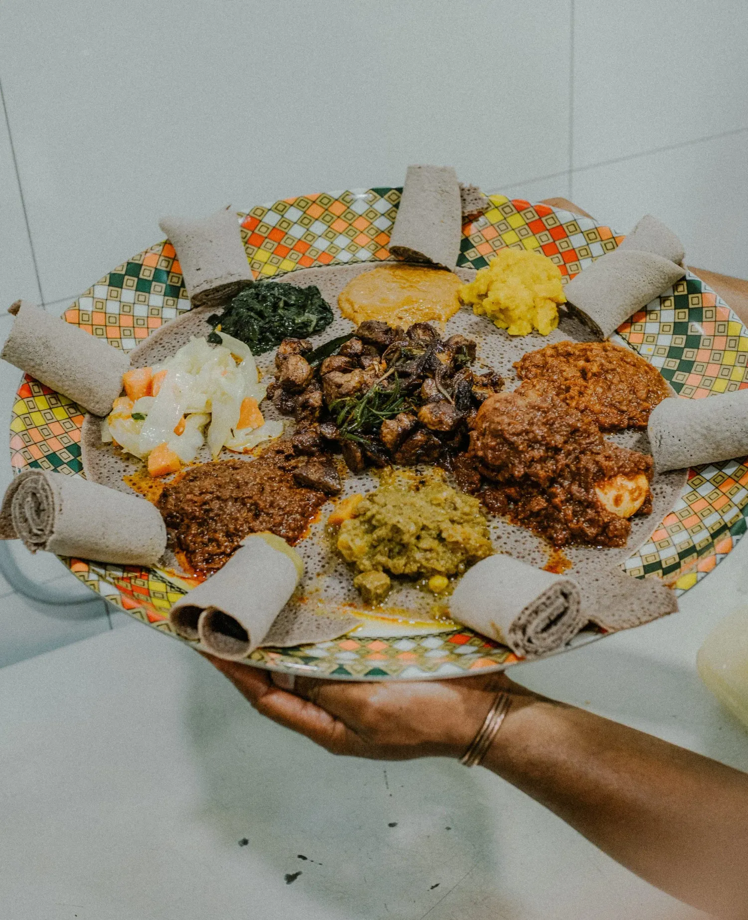 Hand holding a large colorful Ethiopian injera platter filled with assorted stews and rolled injera.