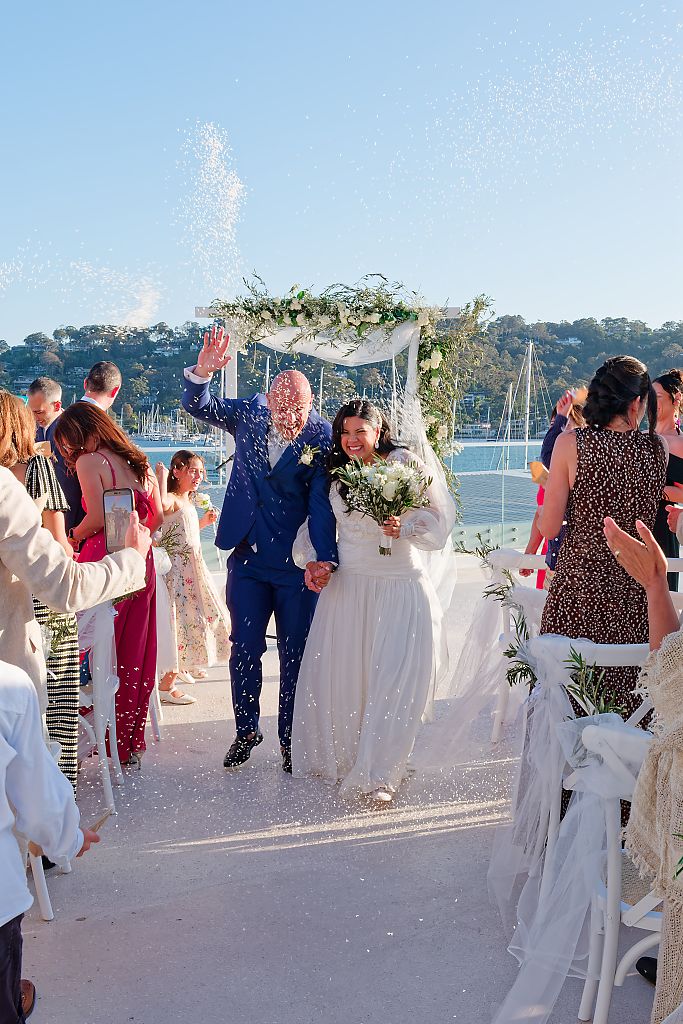 Newlyweds in a blue suit and white gown walk down the aisle through confetti under a floral arch at a waterfront wedding ceremony.