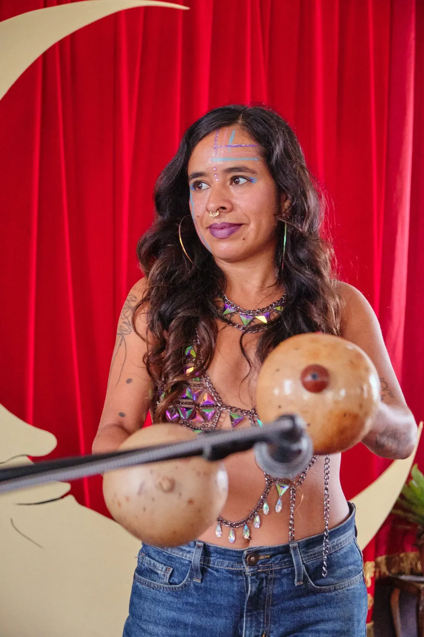 Female performer holding gourd maracas wearing colorful beaded costume with face gems and purple lipstick.
