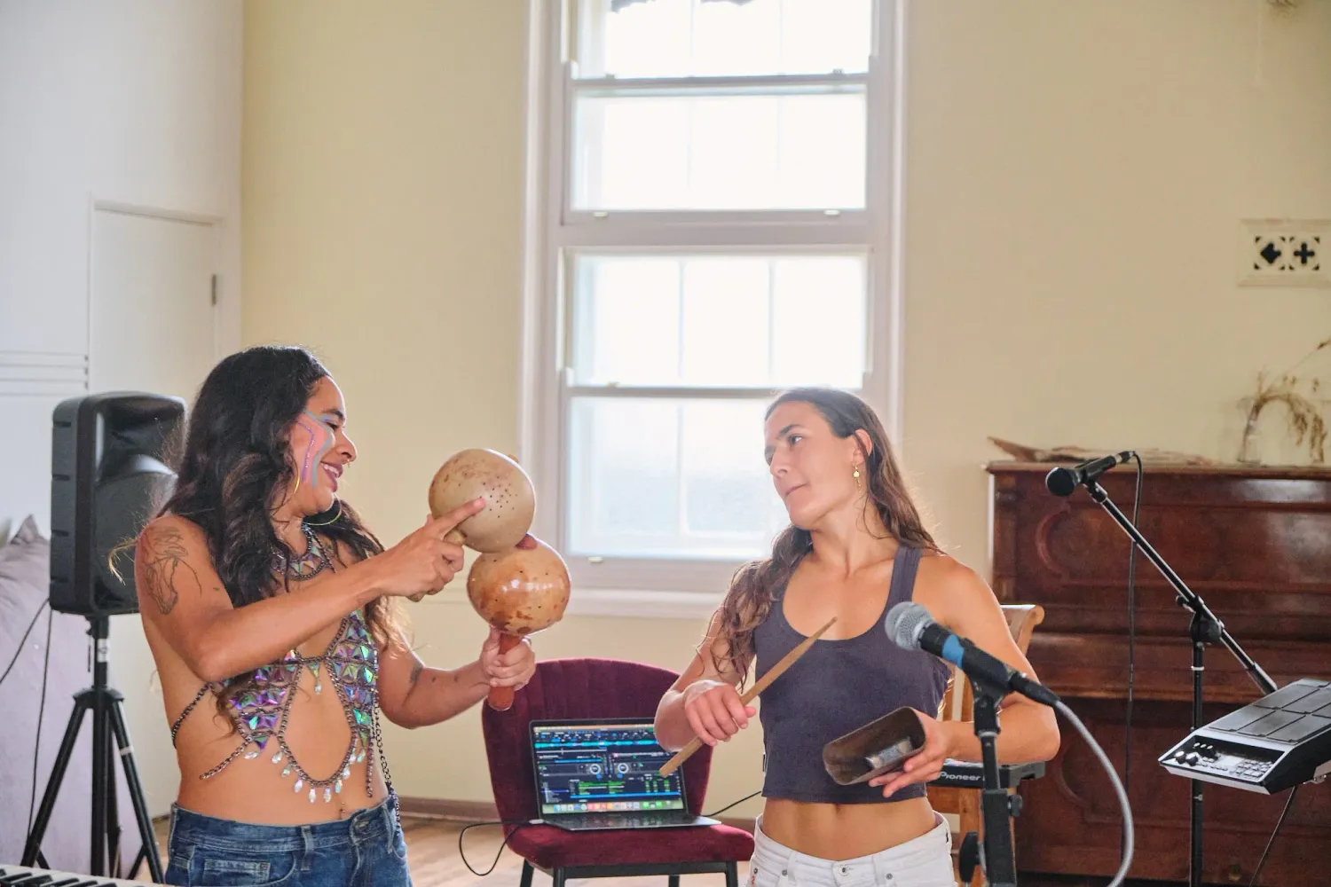Two women smile while playing gourd percussion instruments together in a sunlit music room.