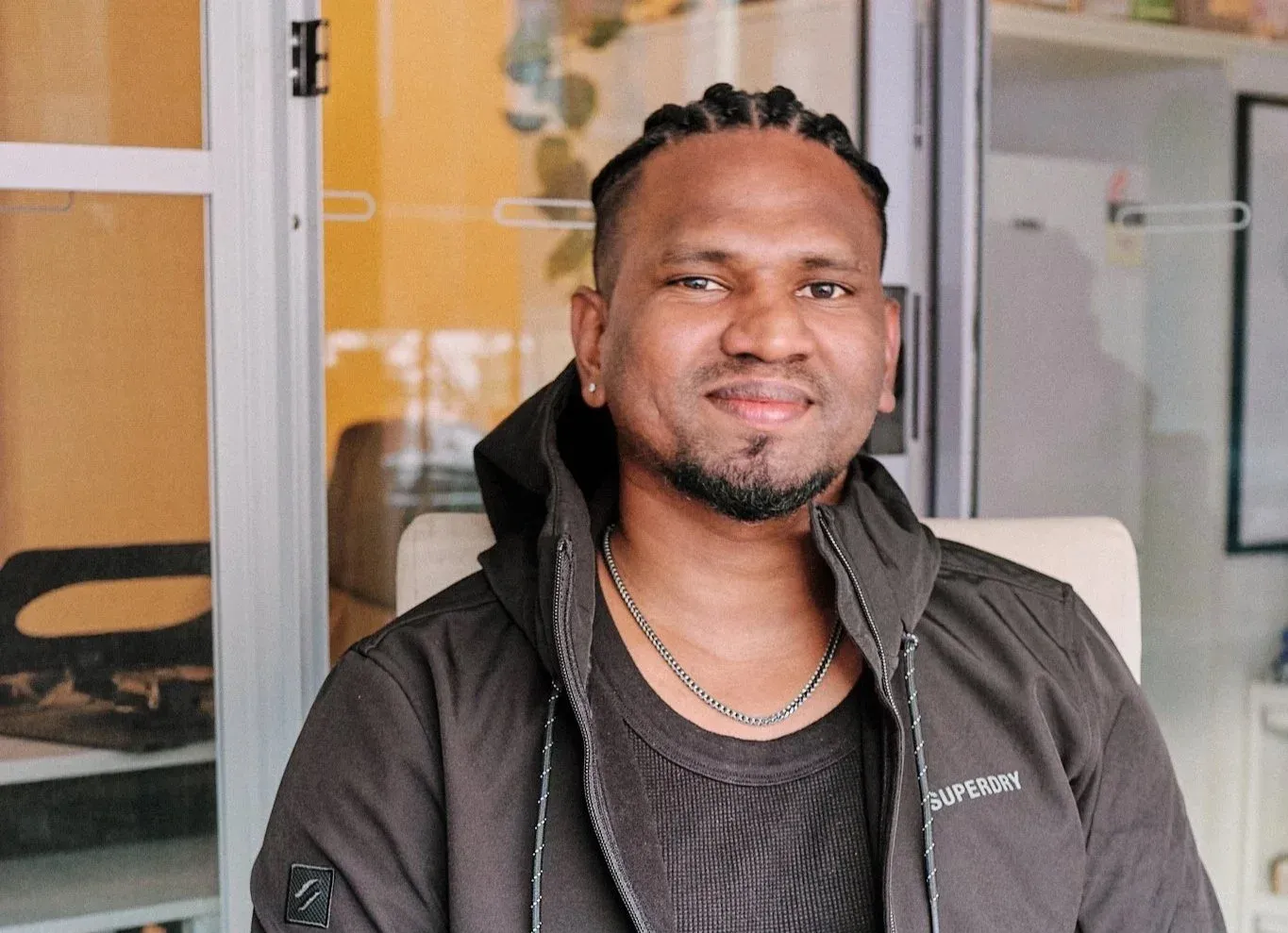 Smiling man with braids in a dark jacket posing indoors in a well-lit space.