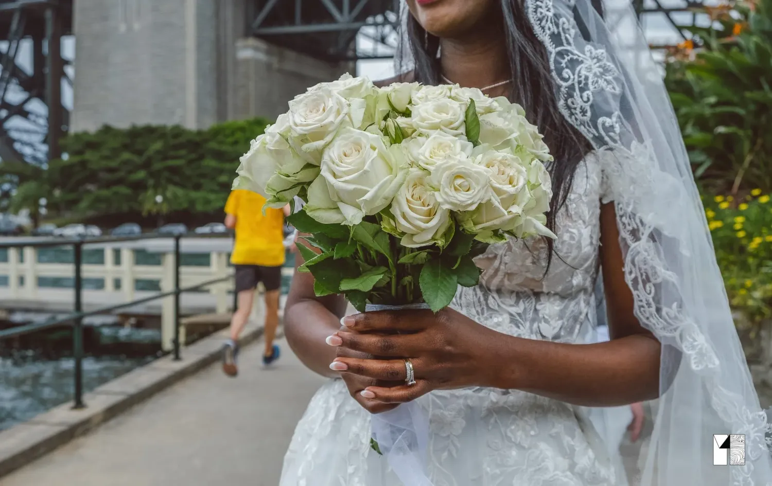 Bride in lace wedding dress holding white rose bouquet near waterfront bridge.
