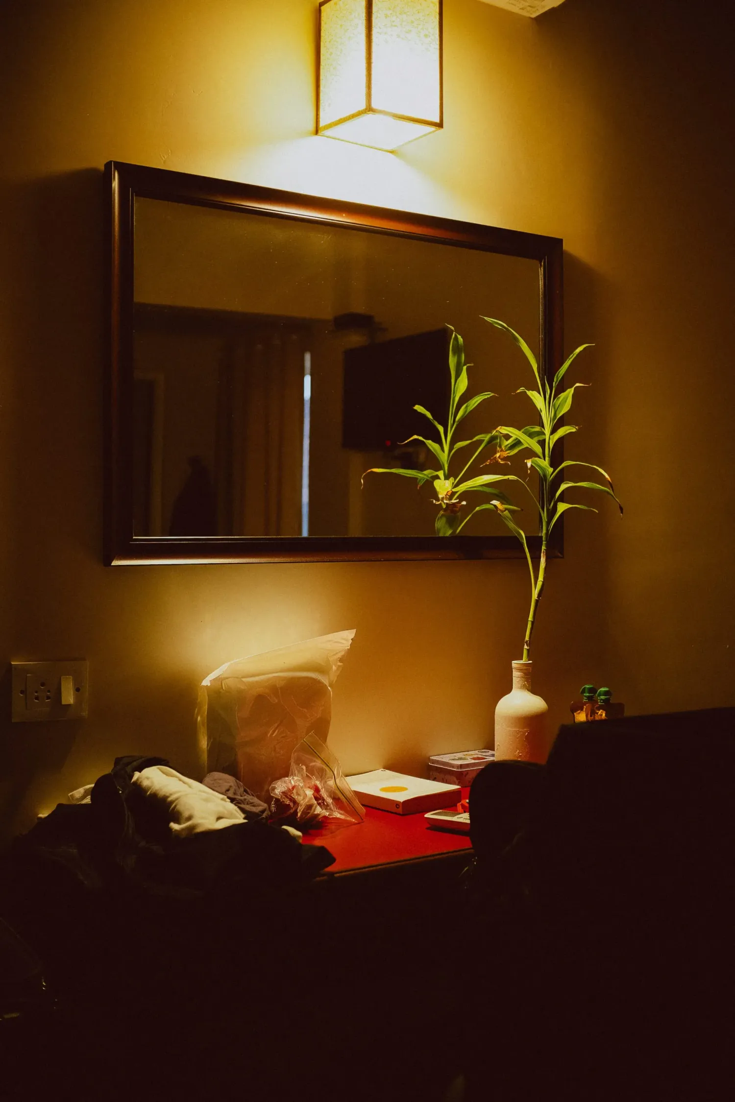 Cozy warmly lit hotel room desk with mirror, plant in vase and wall sconce light.