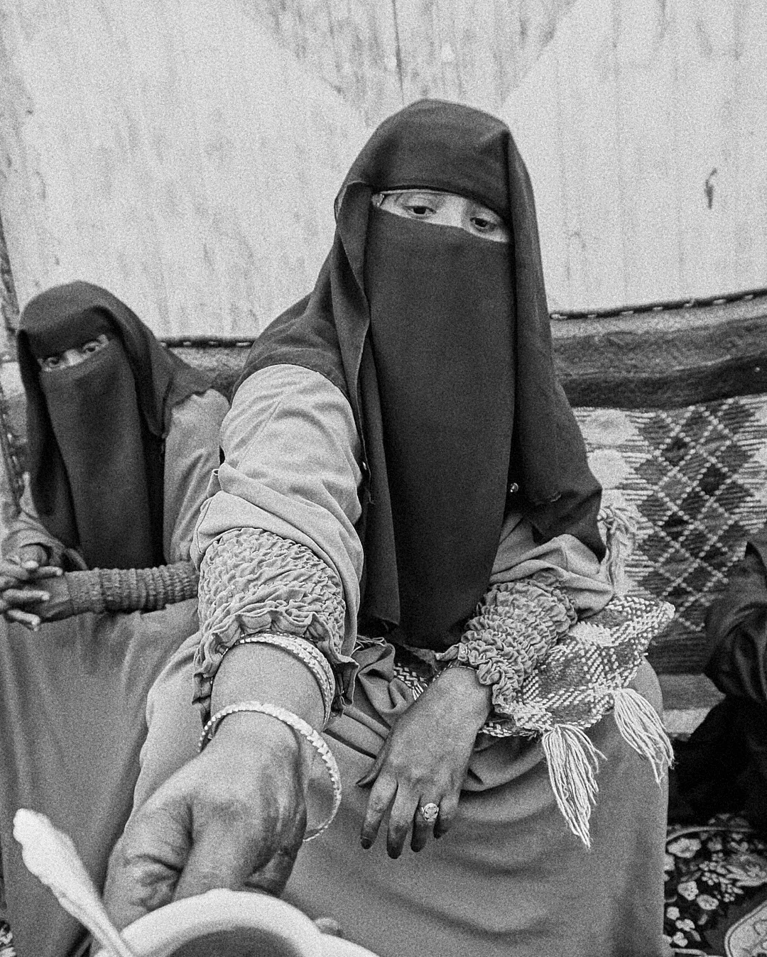 Black and white portrait of two women wearing traditional niqab veils seated against a worn wall.