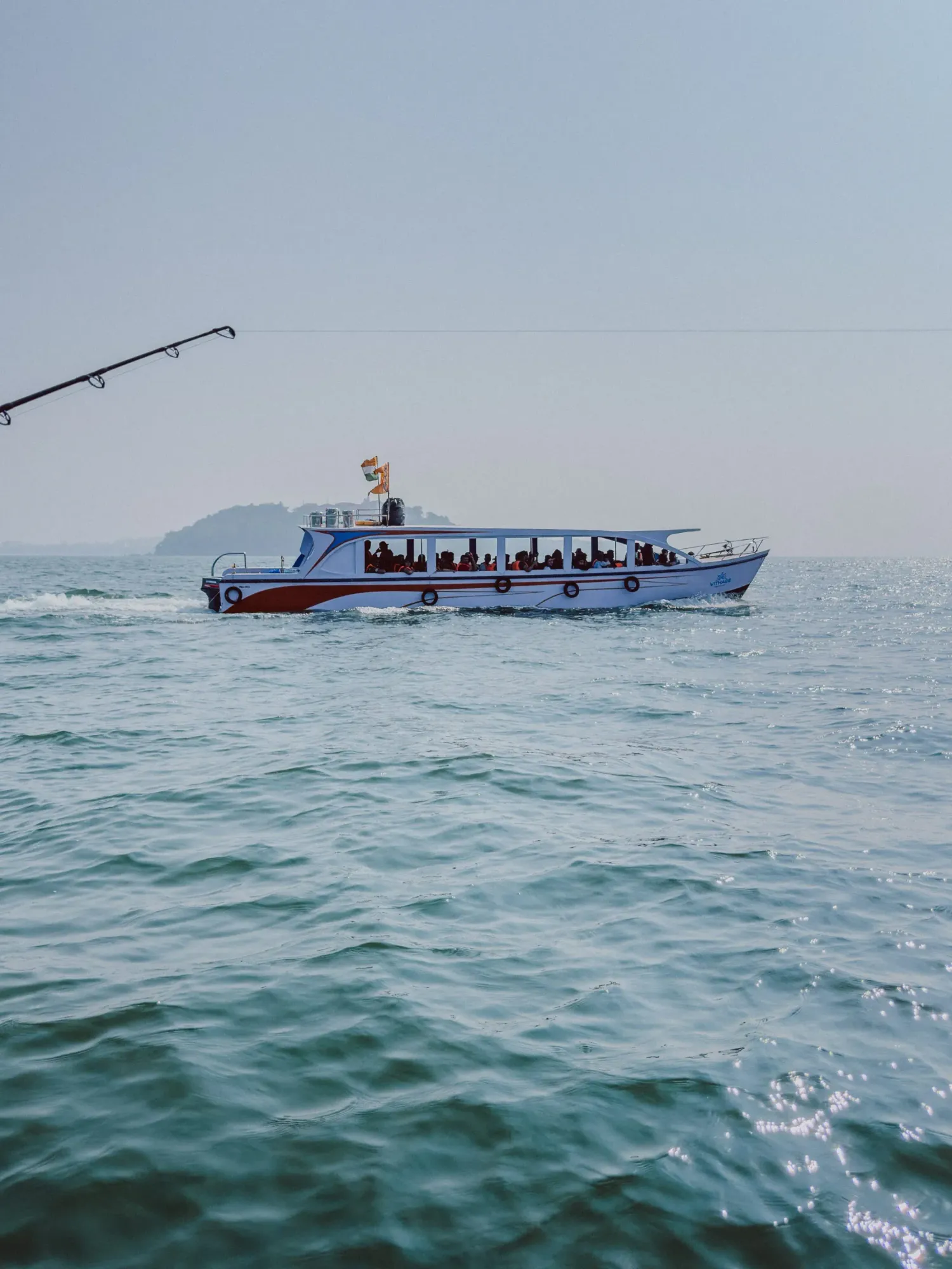A passenger ferry boat cruising on calm turquoise ocean waters with a fishing rod visible in the foreground.