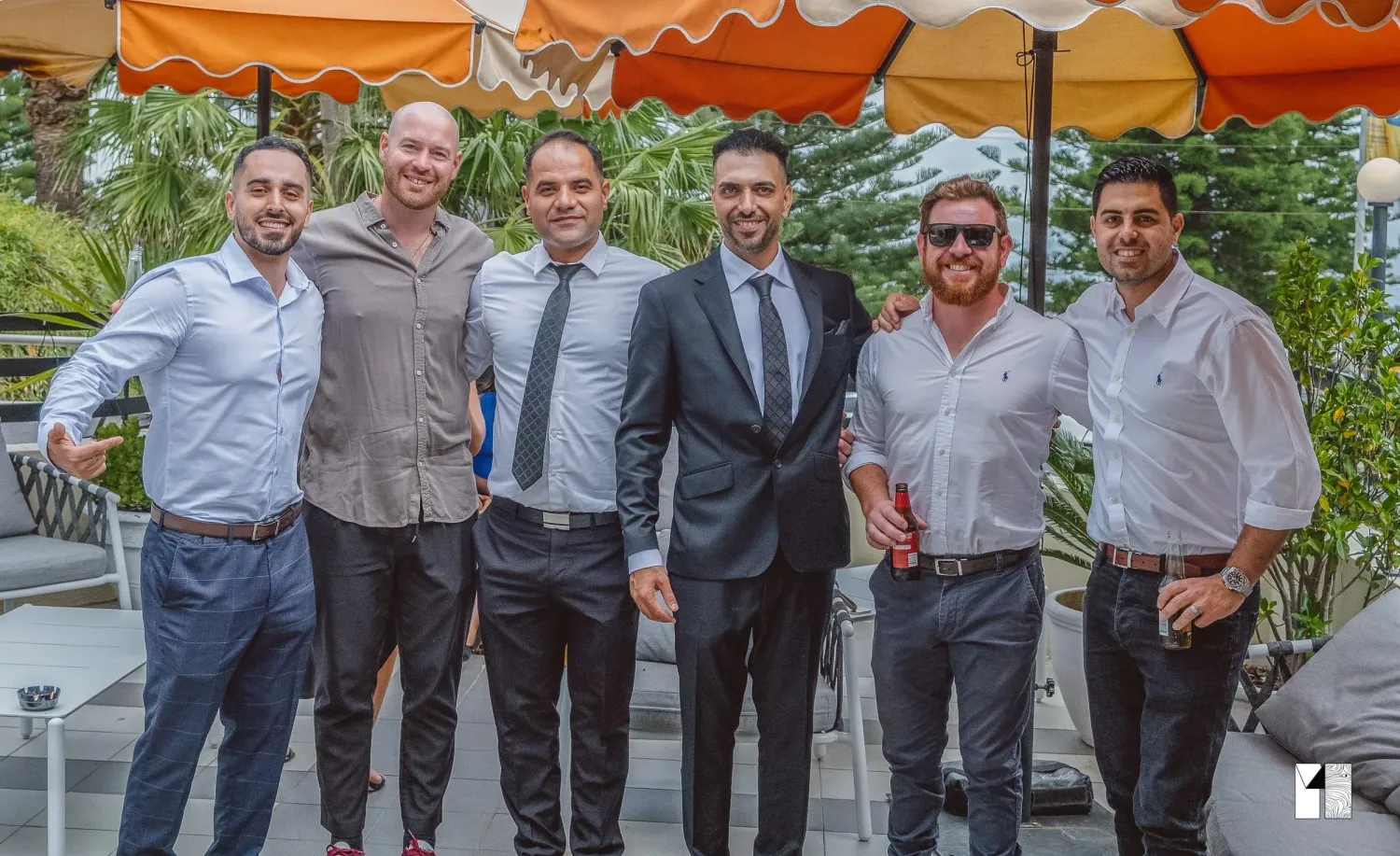 Six men in formal attire posing together outdoors at a wedding celebration event.