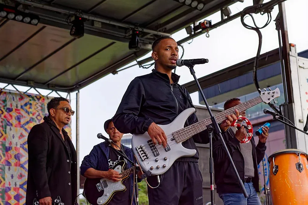 Band performing live on outdoor stage with bassist in foreground at festival.