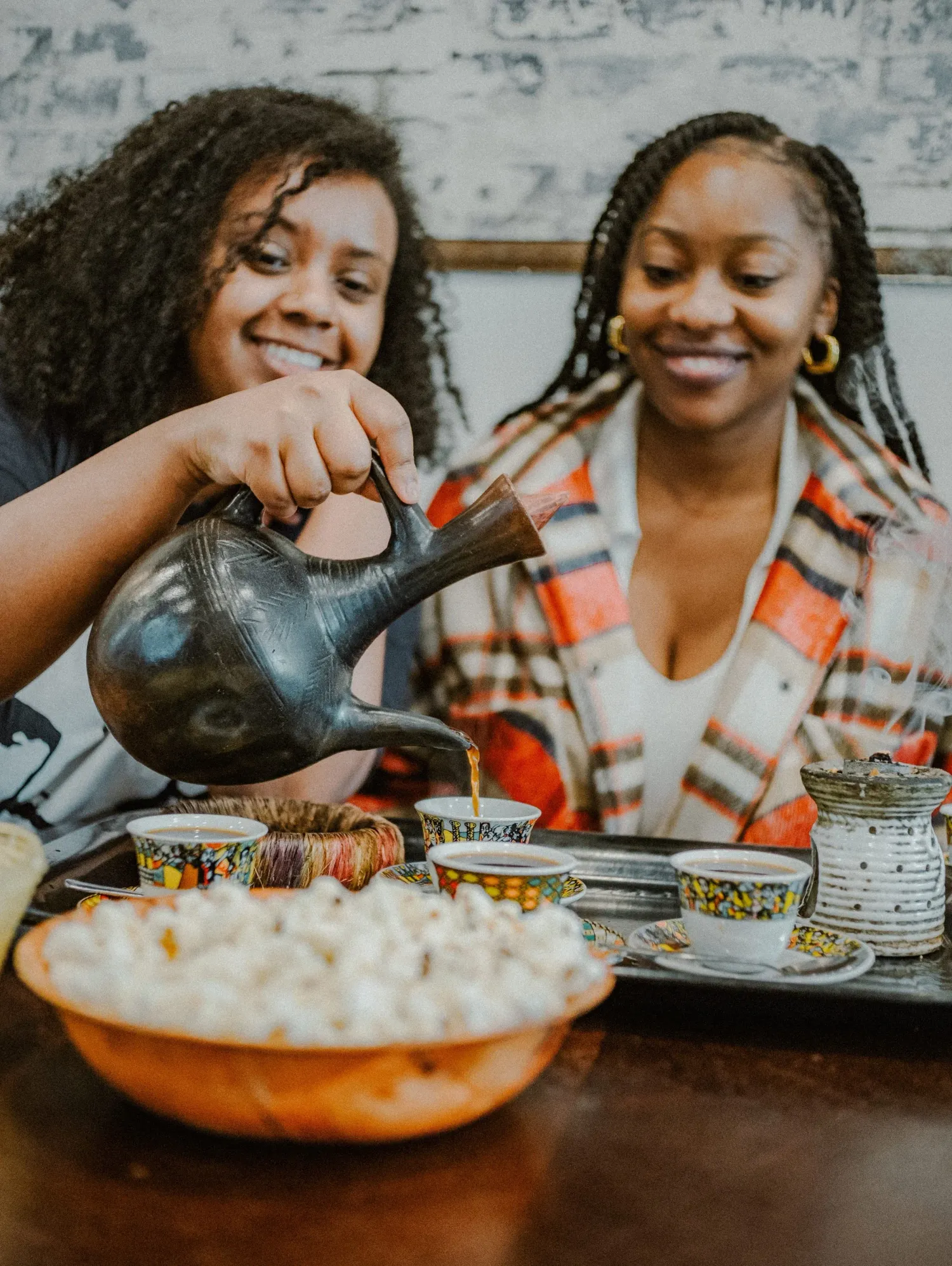 Two smiling women enjoy an Ethiopian coffee ceremony, with one pouring from a traditional black clay jebena pot.