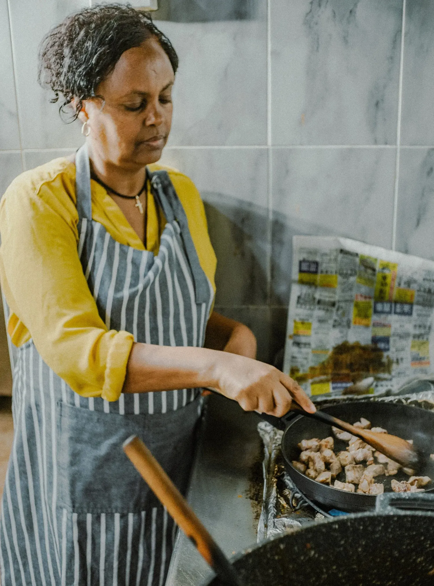 Ethiopian woman in yellow shirt and striped apron cooking meat in a large pan in a kitchen.