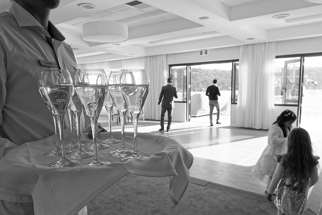 Server holding a tray of champagne flutes in a bright event space as guests walk toward a terrace with large windows.
