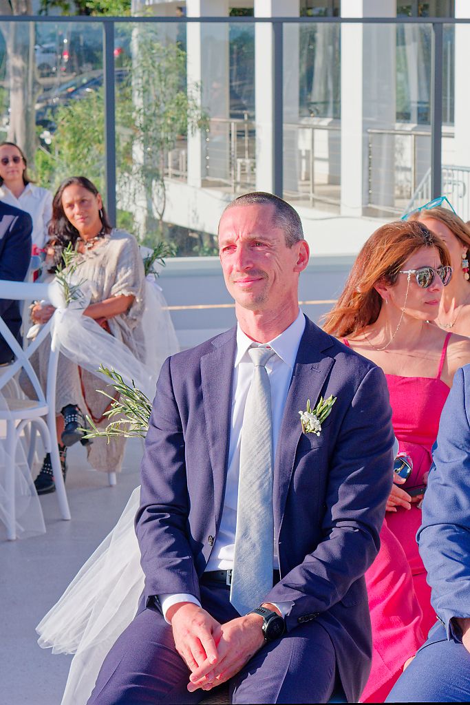 A man in a navy suit with a boutonniere sits with hands clasped among guests at a sunny rooftop wedding ceremony.