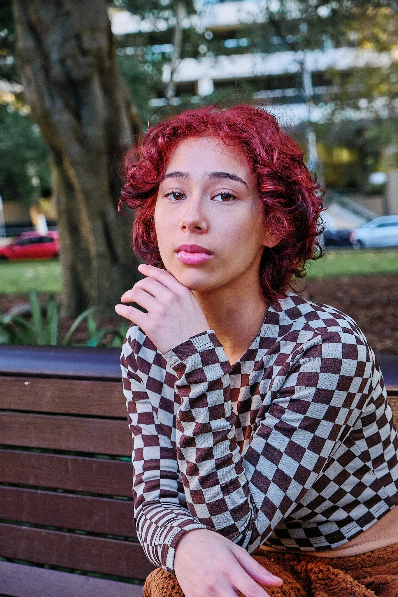 Woman with red curly hair in checkered top sitting on park bench looking at camera.