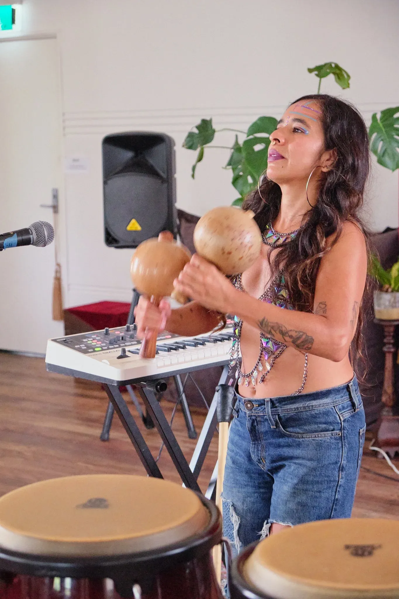 Female performer shaking gourd maracas near conga drums with keyboard and speaker in background.