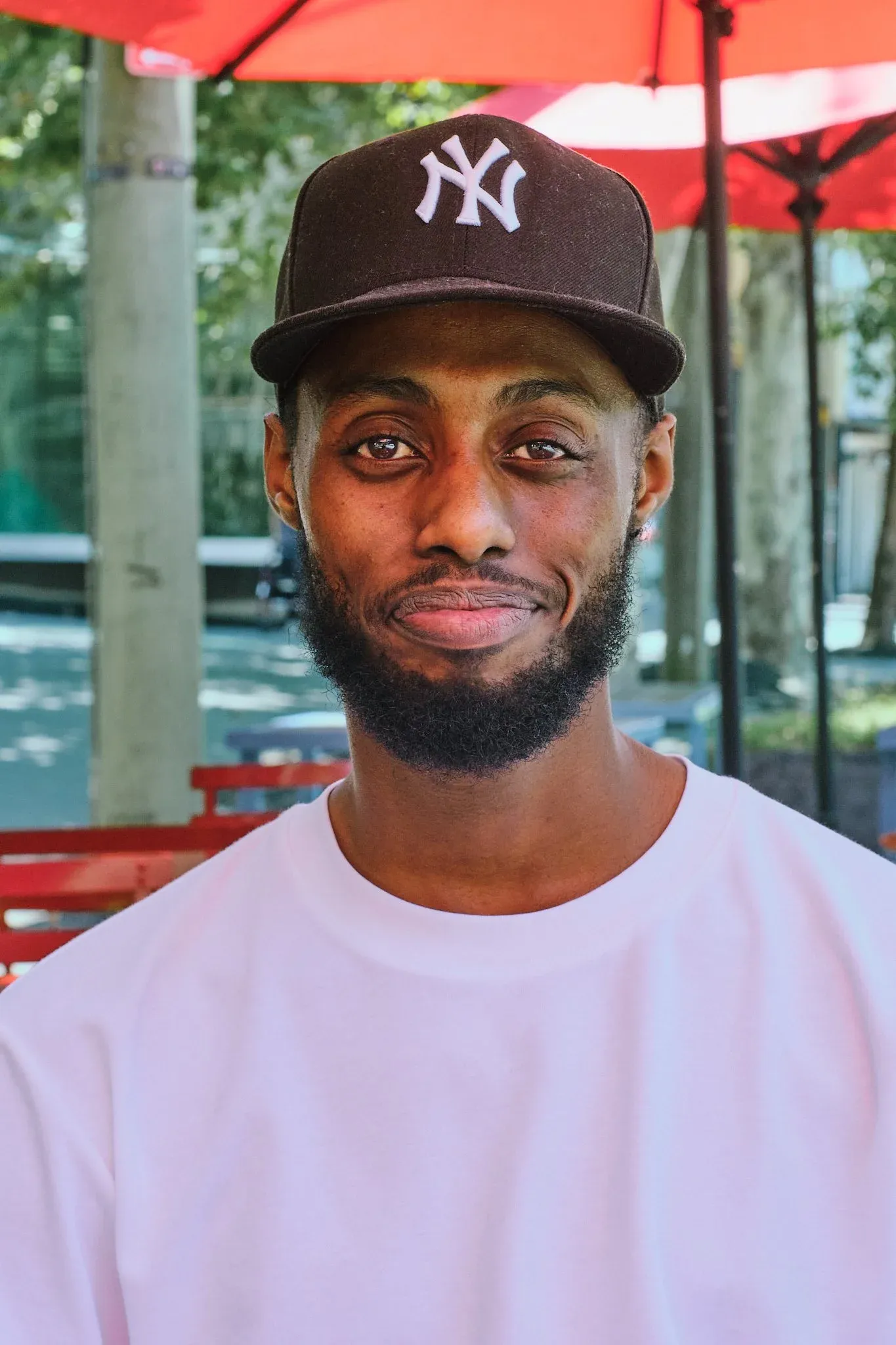 Young Black man smiling wearing NY Yankees brown cap and white tee under red umbrella outdoors.