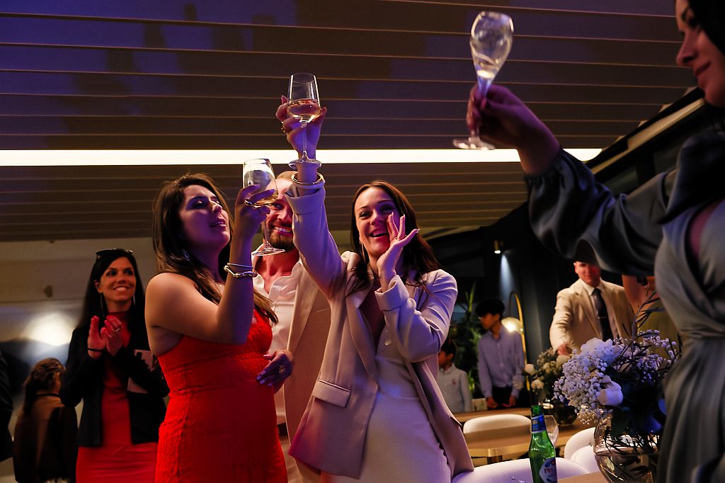 Wedding guests in formal attire raise champagne glasses in a toast at an indoor reception while others applaud and smile.