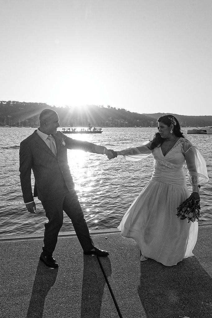 Black and white photo of a bride and groom holding hands and dancing on a waterfront promenade with the sun glowing over the water.