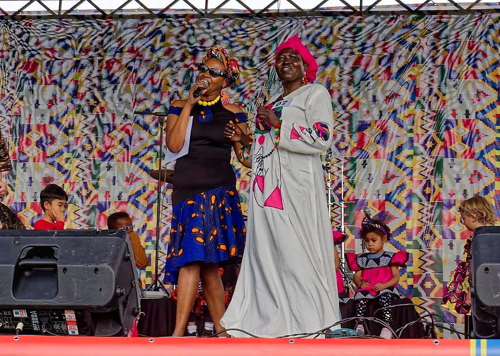 Two women perform on stage, one in a blue floral skirt and one in a flowing white dress.